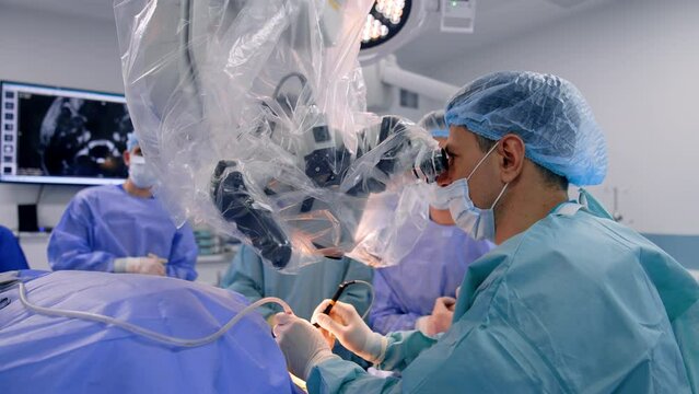 Focused neurosurgeon operates patient looking attentively into microscope. Doctor in mask talks to his colleagues.