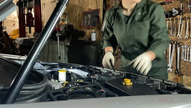 Young woman in coveralls works at a car service station conducting technical car maintenance indoors. Female mechanic replacing engine oil, inserting a funnel into oil filler neck. Tools on background