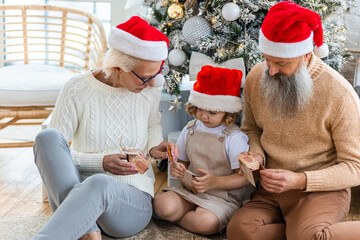 Attractive senior grandparents doing Christmas and New Year advent calendar with granddaughter, cute little girl. Looking happy and excited sitting near decorated tree in red Santa hats