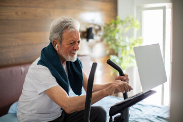 Senior Man with Earphones Using a Stationary Exercise Bike While Looking at a Laptop at Home - obrazy, fototapety, plakaty