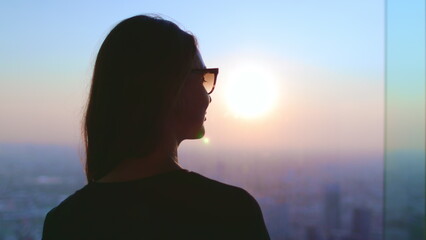 Tourist woman watching sunset over cityscape. Girl silhouette in glasses looking in window, colorful sky, city buildings, aerial view. Baiyoke Tower Sky, Thailand. Travel, tourism