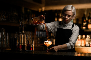 Female bartender pours an iced cocktail from a mixing glass into a cocktail glass