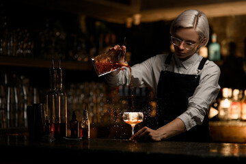 Female bartender pours an iced cocktail from a mixing glass into a cocktail glass with a large piece of ice
