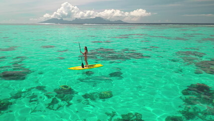 Woman standing on yellow sup board with paddle. Crystal turquoise water coral reef, tropical...