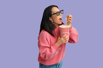 Young Asian woman in 3D glasses with bucket of popcorn on purple background