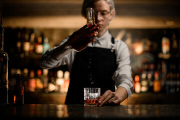 Female bartender holds a small geyser bottle and pours the drink in small portions into a glass with ice