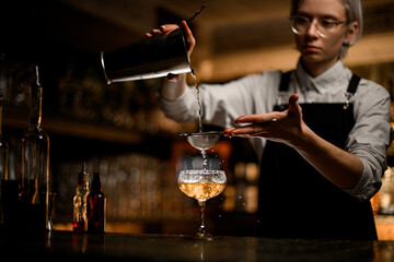 Female bartender pours a clear liquid from a metal beaker into a glass with a sieve