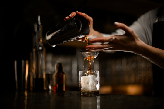 Female hand pours a cocktail through a strainer from a metal tumbler into a glass tumbler