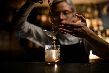 In the foreground is a glass with a piece of ice into which a cocktail is poured through a strainer