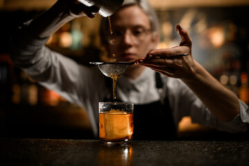 Female bartender pours the last drops of a cocktail from a metal glass into a glass with ice