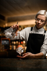 bartender in glasses pours the last drops of clear drink into a stemmed glass with a large square...