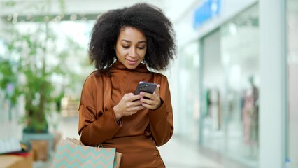 Young beautiful happy woman shopper joyful customer stand in shopping center mall and uses a mobile phone browsing chatting typing smartphone Satisfied glad with gift color package bag in store