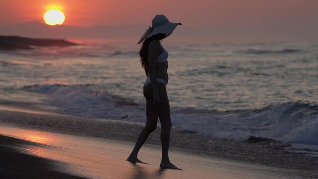Woman tourist in white bikini and straw sun hat walking on sandy beach at sunrise on summer morning. Full length female during weekend beach holiday at golden hour. Slow motion, cinematic, handheld