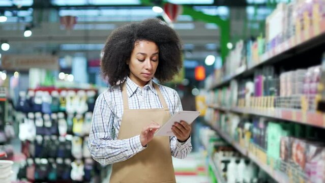 Portrait of a young African American woman supermarket manager in apron with a digital tablet in her hands. Confident female clerk worker in a grocery store or hypermarket saleswoman look at amera