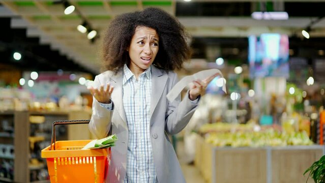 Woman shopper takes out a paper check in a grocery store in supermarket is very surprised at the high prices, rising inflation. Gets shocked looking at check after shopping. consumer standing in mall 