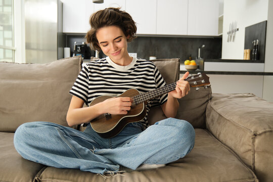 Portrait of cute young woman sitting on sofa, learns how to play ukulele, holding her music instrument, picking chords, resting in living room