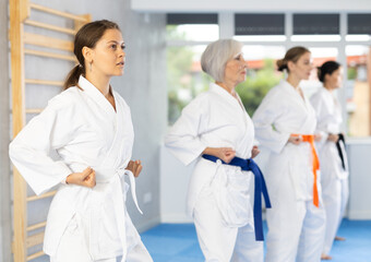 Fototapeta premium Concentrated young girl in white kimono practicing punches in gym during martial arts workout with female group. Shadow fight, combat sports training concept