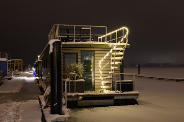 Houseboat with festive lighting moored at the pier covered with snow in winter.Christmas romance afloat: houseboat adorned with festive lights, moored at a snow-covered pier on Schlei, Schleswig.