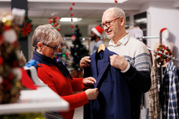 Elderly couple looking at blazers in retail store, searching for formal clothing to wear on christmas dinner celebration. Customers checking fabric of suit jacket for elegant outfit, festive decor. - obrazy, fototapety, plakaty