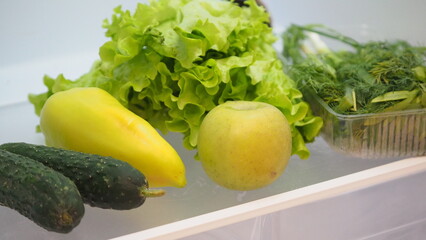 Woman hand taking, grabbing or picks up fresh fruits, vegetables and herbs out of open refrigerator shelf or fridge drawer full of fruits, vegetables, apple, tomatoes, cucumbers, lettuce. Healthy food