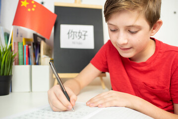 An 11 year old child is learning Chinese. a happy European boy sits at a desk, in front of him is a Chinese flag, on the desk is the hieroglyph “Hello” in Chinese, the boy is doing his homework
