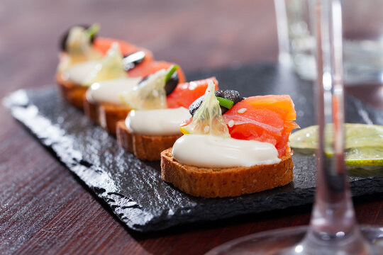 Canapes on toasted bread with smoked salmon, olives and creamy sauce on black serving board..
