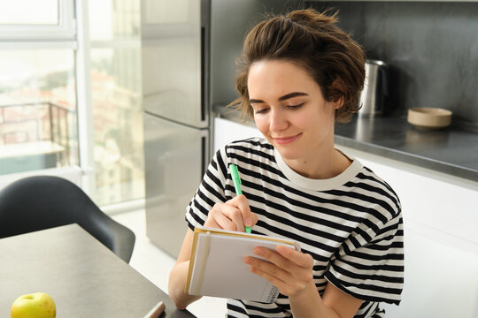 Close up portrait of smiling, cute woman with notebook, writing down recipe, making notes or grocery list for shopping, sitting in the kitchen