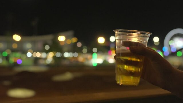 This video shows a close up view of an anonymous hand toasting a beer to a night time event.
