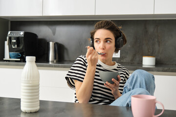 Cute modern woman, student eating quick breakfast, having cereals with milk and coffee, listening to podcast or music in wireless headphones