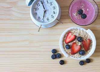 flat lay of  alarm clock, breakfast with a  glass of blueberry smoothie and oat or granola in white bowl, fresh blueberries, strawberries on wooden table. Healthy breakfast concept.