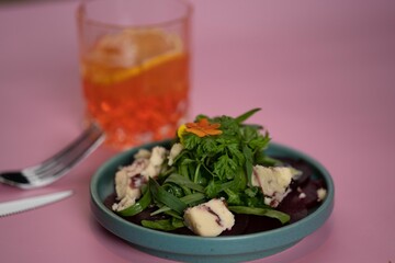 Closeup of a bowl of beetroot salad with cheese and greens with a drink on a pink background