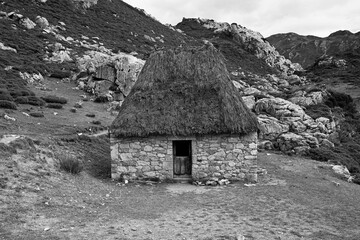 a small thatched roof stone hut on a hillside with mountain range in the background