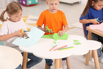 Cute little children cutting color paper with scissors at desks in kindergarten. Playtime activities