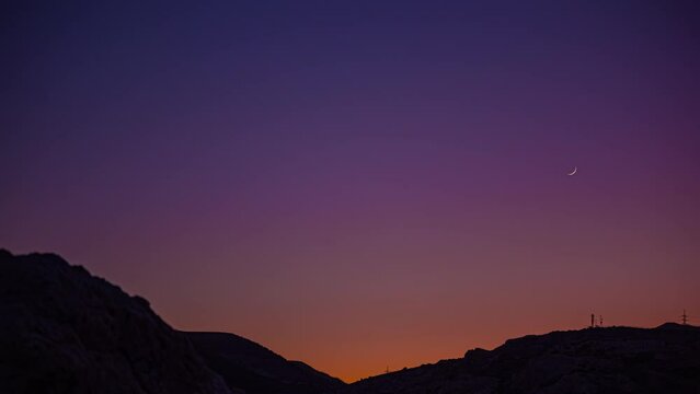 Low angle shot of moon movement in timelapse on colorful night sky after sunset over hilly terrain.