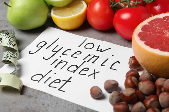 Paper with words Low Glycemic Index Diet, measuring tape and food on grey table, closeup