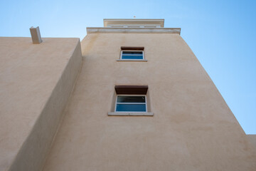 Looking up at the tower of a southwestern style building with blue sky overhead. The building has two small windows and is cream colored.