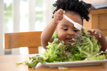 Cute little African girl likes to eat green vegetables, use her hands to pick up lettuce and put it in her mouth - obrazy, fototapety, plakaty