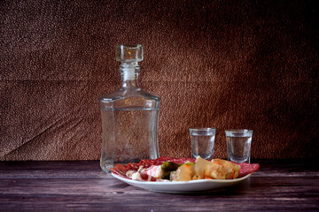 A plate with a variety of snacks on a wooden table, next to a glass decanter of cold vodka and two...