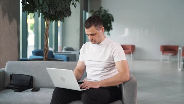 close-up portrait of a young man in a white T-shirt against the backdrop of a business center, tiredly sitting behind a laptop, working, and nervously finishing the task