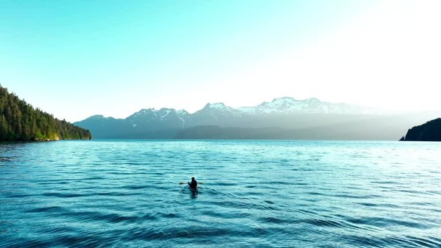 Aerial of a man in a small canoe boat sailing in the ocean in Alaska with mountains in background