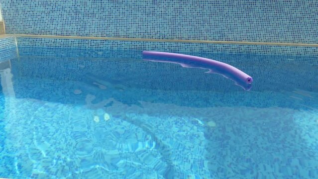 A clip of a blue mosaic tile swimming pool. A purple pool noodle is thrown in the water making many ripples in the water. The noodle stays floating in the water.