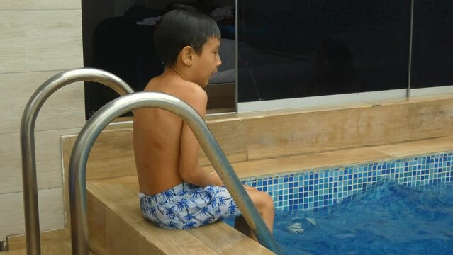 A small young boy sitting wet outside a swimming pool shivering from the cold one feels after getting out of the water.