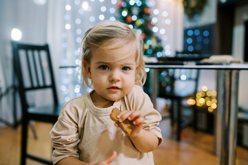 Little girl with a cookie in her hand stands near the table on the background of the Christmas tree