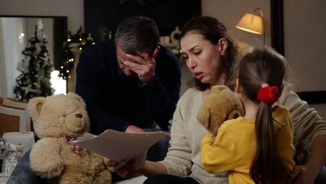 Sad middle-aged couple with a child looking paper documents with tax debt, mortgages or loans, utility bills. Crisis and financial problems during the Christmas holidays