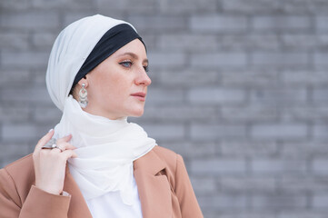 Portrait of a young blue-eyed woman in a hijab against a gray brick wall. 