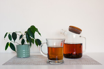 Black tea in a glass cup and glass teapot on a napkin and green plant on white table with blured white background