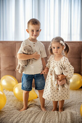 Big brother and his sister are standing on a bed surrounded by balloons and hugging while holding...
