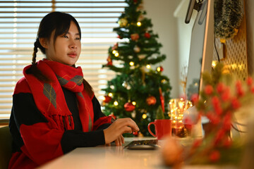 Young woman in sweater looking at computer monitor while working in decorated living room for Christmas
