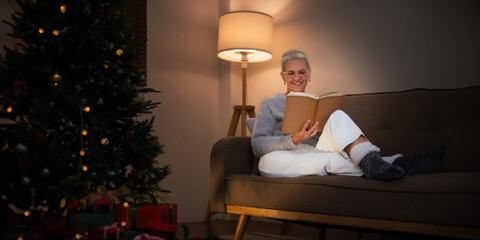 Smiling elderly woman with a cup of tea and a book relax in side of a decorated Christmas tree in...