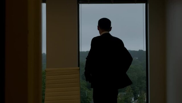 Businessman in a black suit is walking towards a big window that looks at the buildings. Slow motion shot